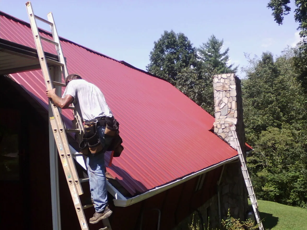 Roofer on a ladder beside a red metal roof and stone chimney — Soffit & Fascia Repair by Roofing Brothers in Port Royal